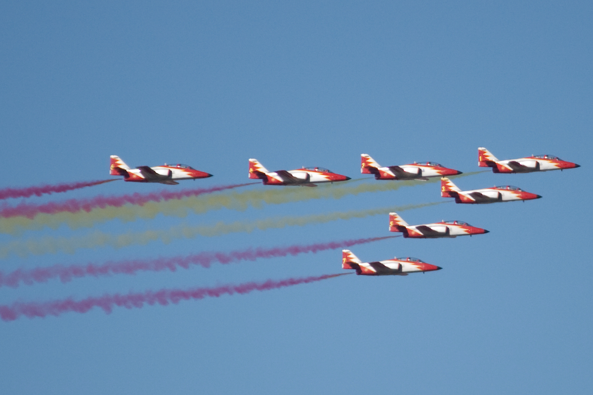 Group of jets flying in a triangular formation emitting streams of red and yellow smoke.