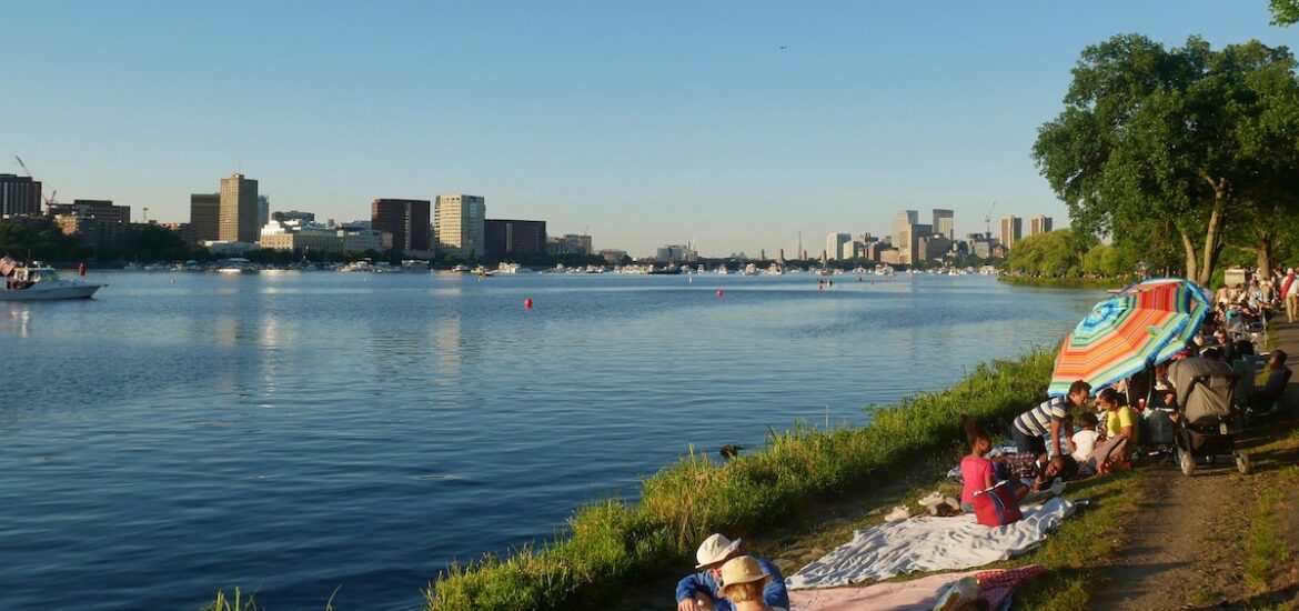 Lots of people line the bank of the Charles River sitting on blankets and enjoying a great July 4th picnic spot in Boston