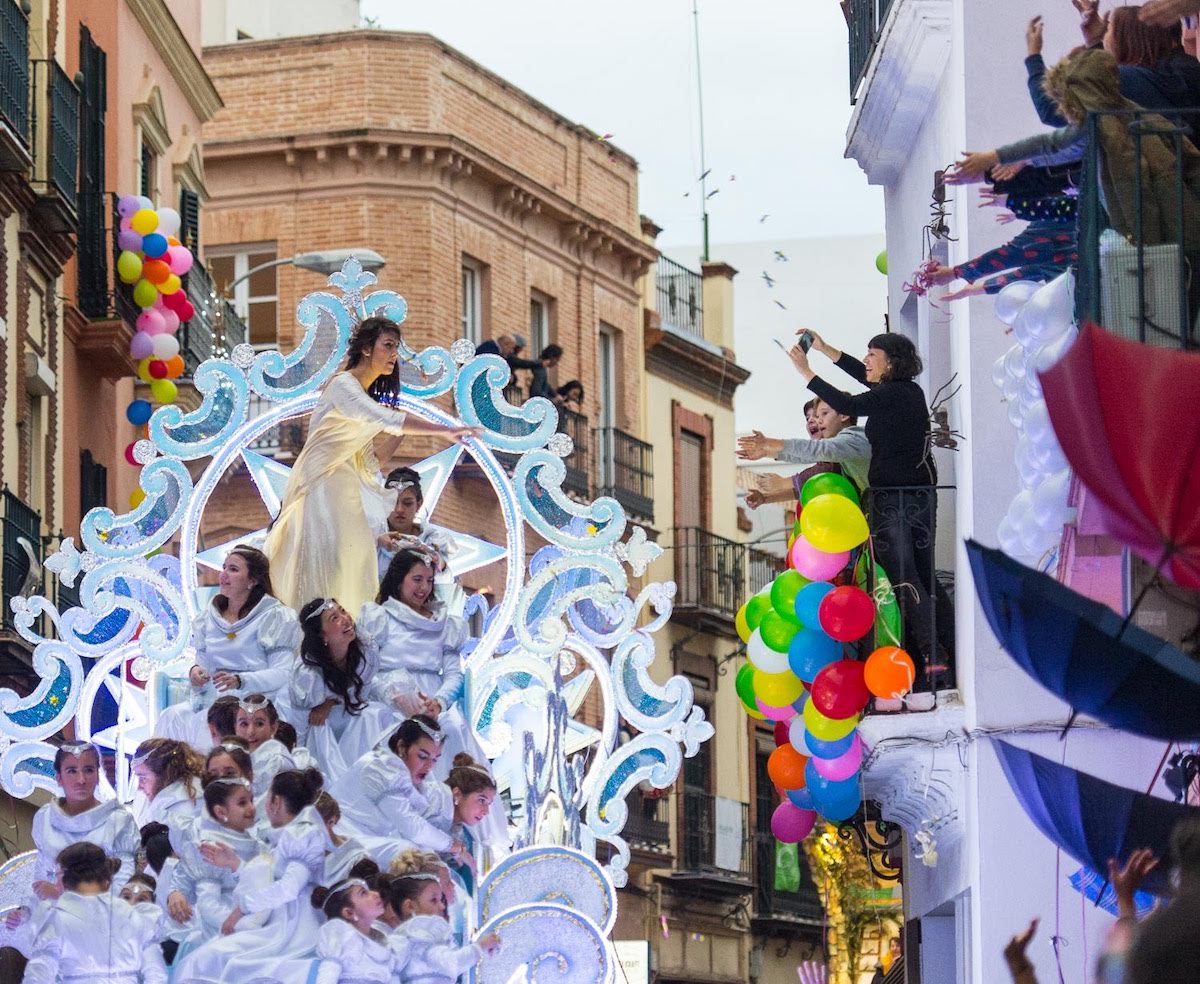 People watching a parade from a balcony as a large float full of people dressed in white costumes passes by