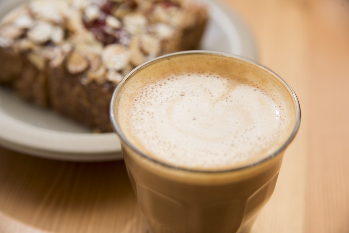 extreme close up of a coffee drink with foamed milk in a clear glass, with a pastry on a white plate in the background