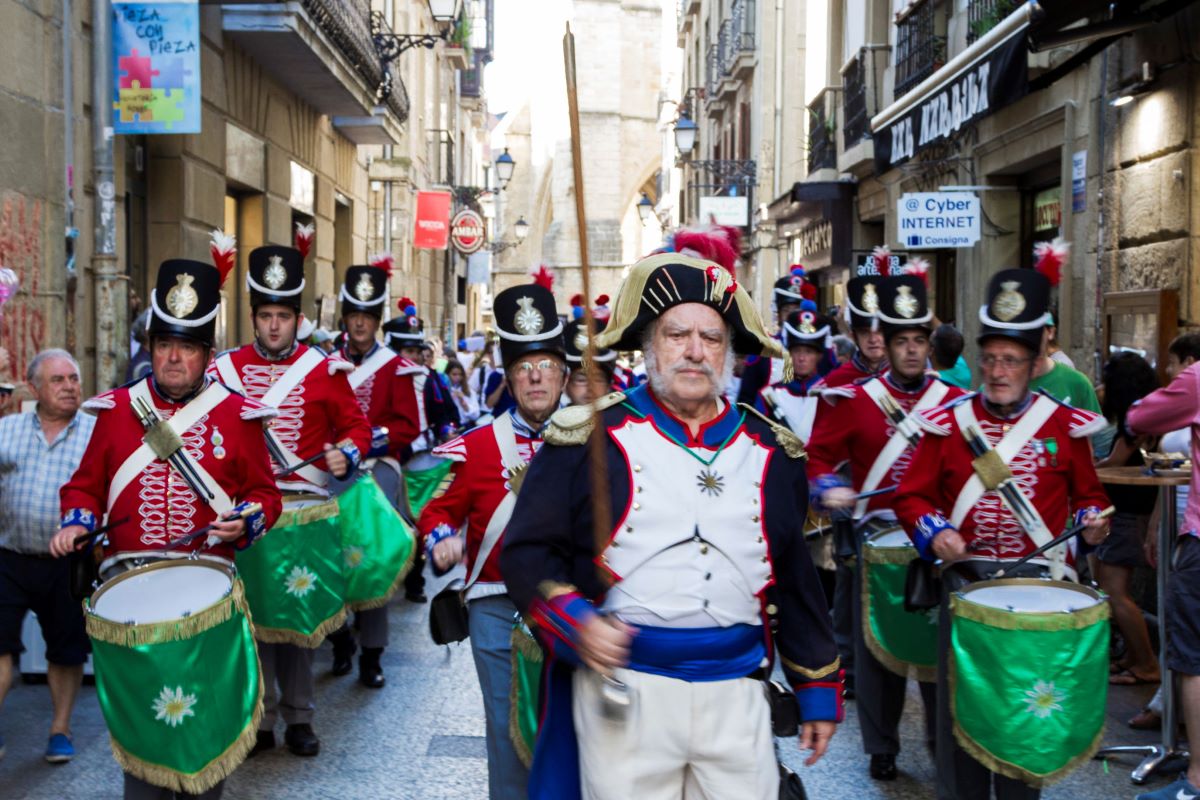 Men dressed in traditional Spanish clothing marching down a street with drummers behind them.