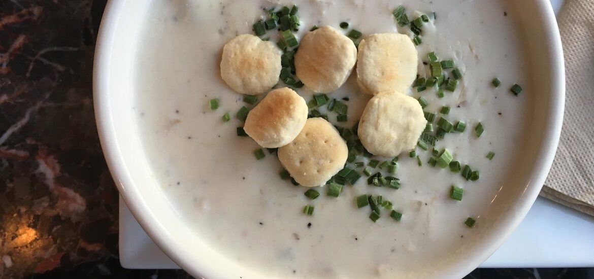 Flat lay shot of a bowl of New England clam chowder topped with chives and oyster crackers on a black marble table