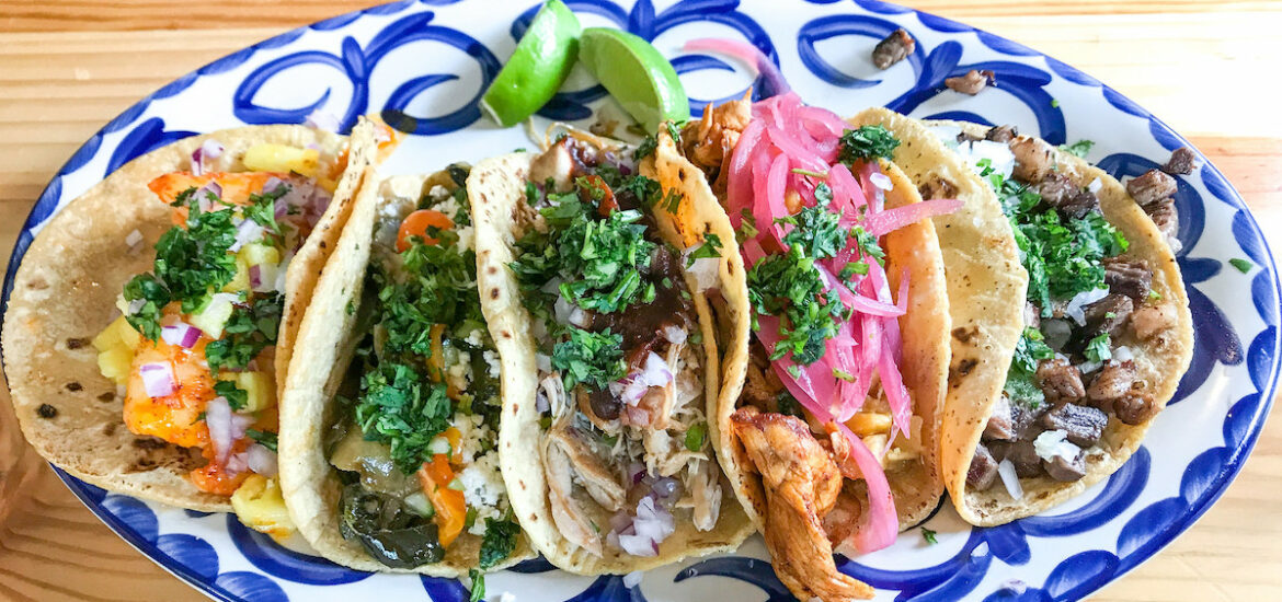 A wooden table with a blue and white painted platter on top of it with five tacos and lime wedges