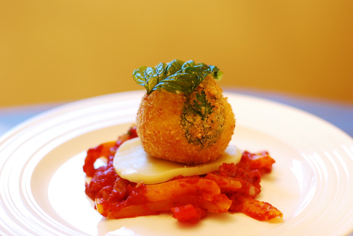 Extreme close up of one arancini, a little ball of risotto breaded and fried, atop chopped tomatoes and a slice of mozzarella cheese