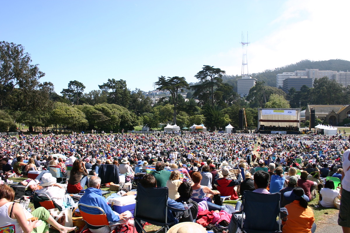 A massive crowd of people sit in lawn chairs spread over a huge lawn, facing a stage production of Opera in the Park in San Francisco