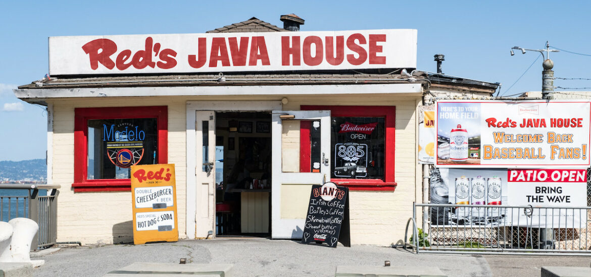 exterior shot of a small white building on a pier with a blue sky. Big red letters spell "Red's JAVA HOUSE" on a sign