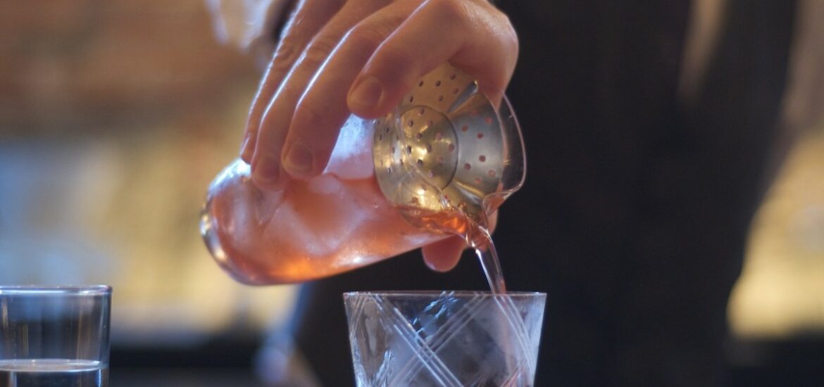 A bartender pours a pale red cocktail from a shaker into a cocktail glass.