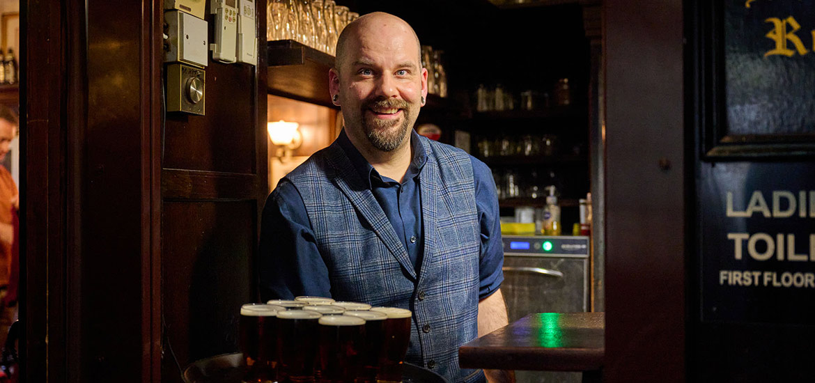 A smiling bartender in a blue shirt and checkered vest stands behind the bar with several pints of beer ready to serve, in a warmly lit pub.