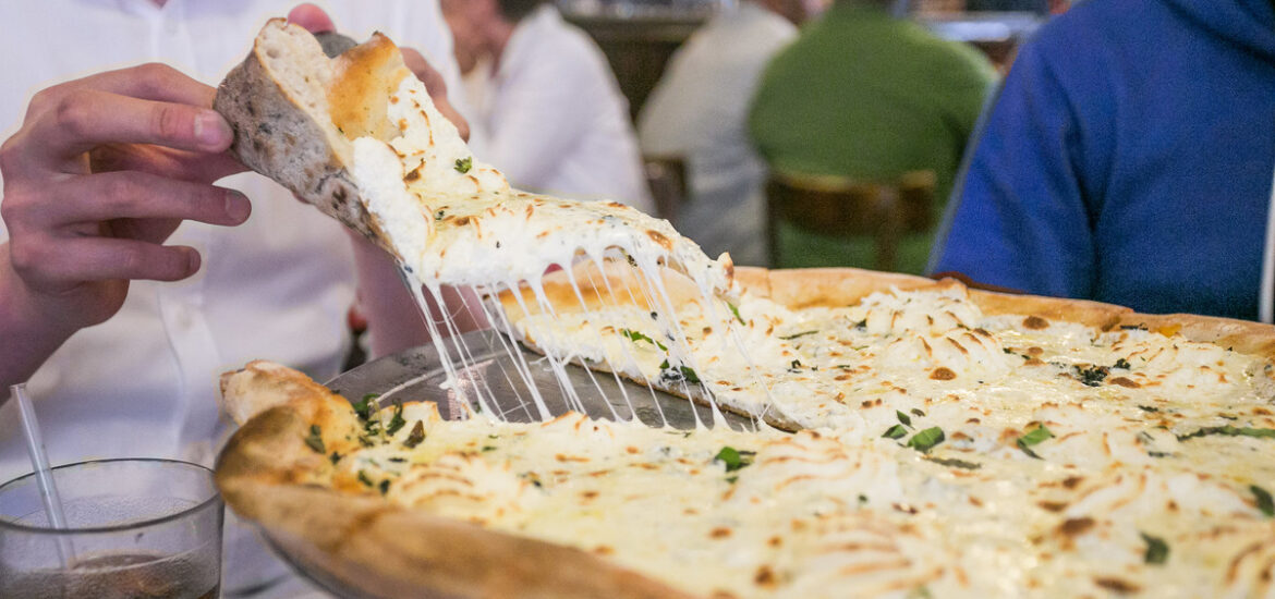 Man pulling a slice of white pizza from a pan with strings of cheese connecting it to the main pizza