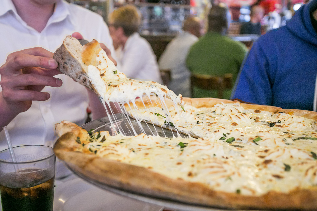 Man pulling a slice of white pizza from a pan with strings of cheese connecting it to the main pizza