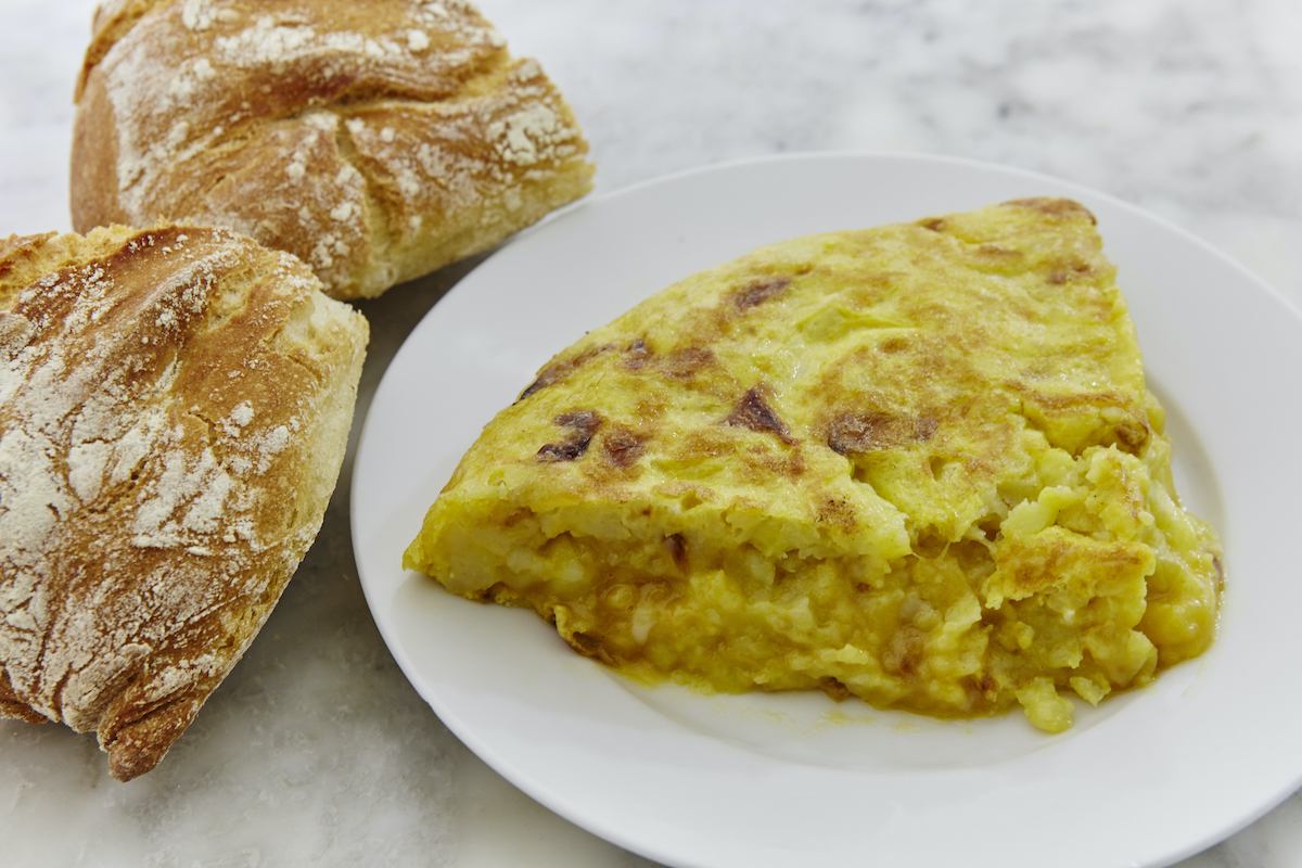 Slice of potato omelet on a white plate beside two bread rolls