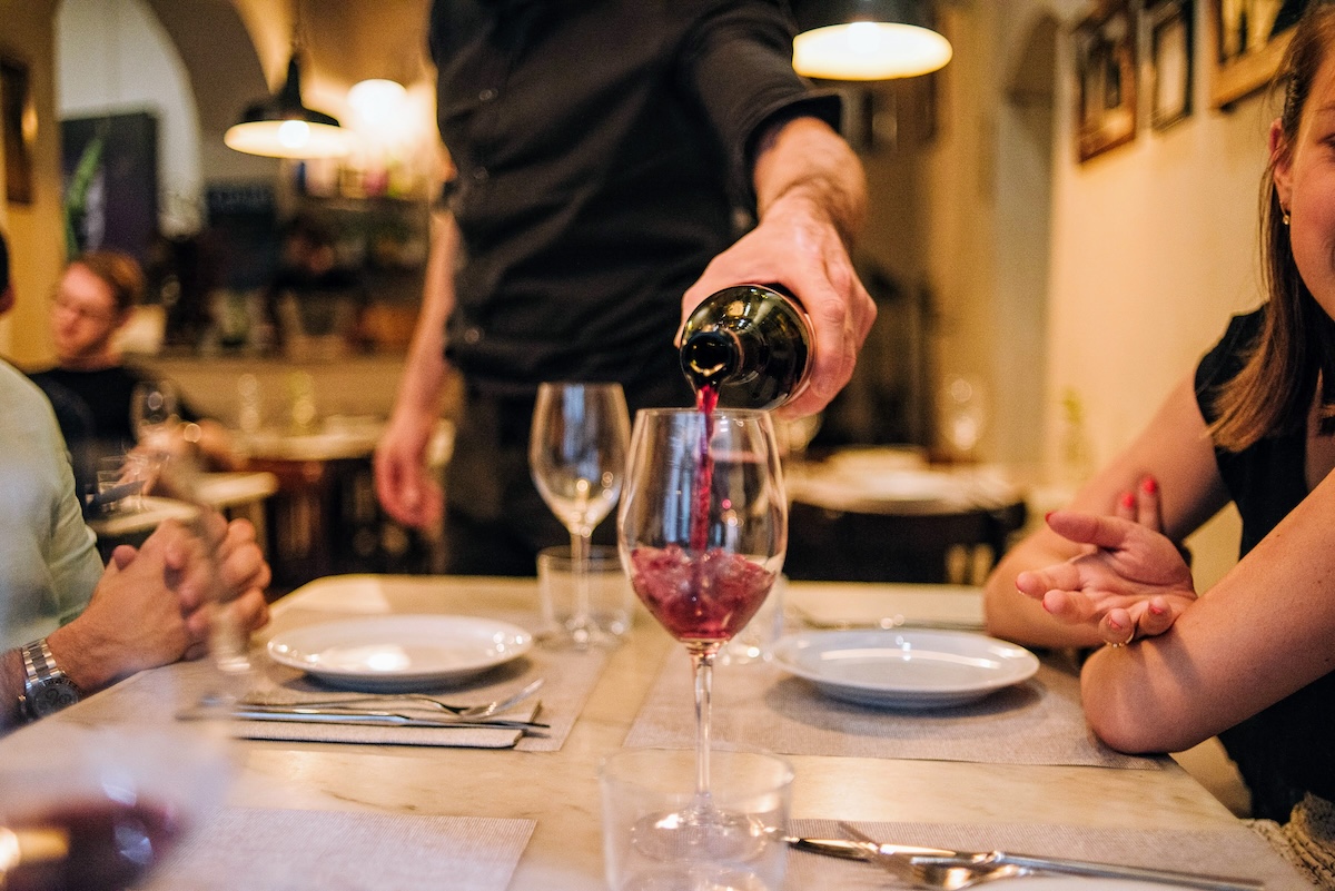 waiter pouring red wine into a wine glass at a restaurant with a group of people around a table