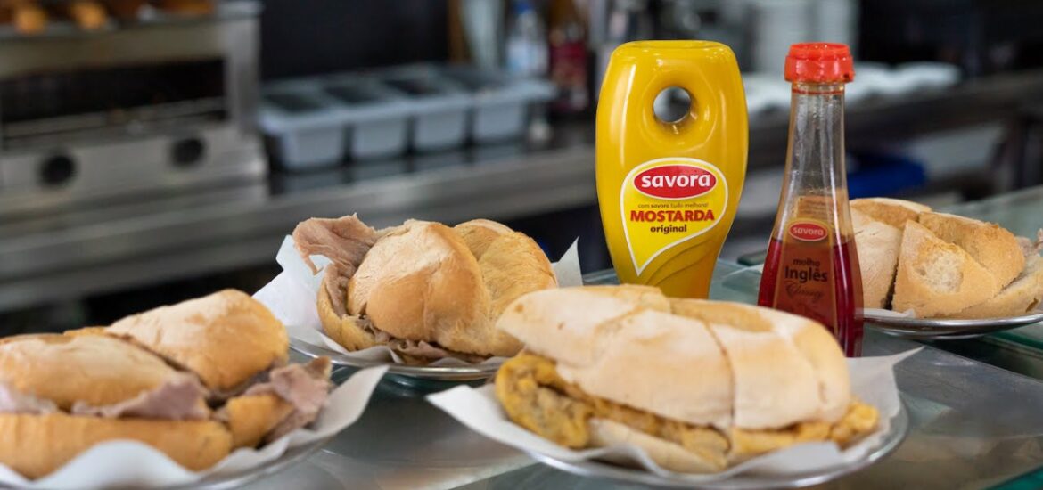 Bifana sandwiches on a restaurant coutner with mustard and other sauces