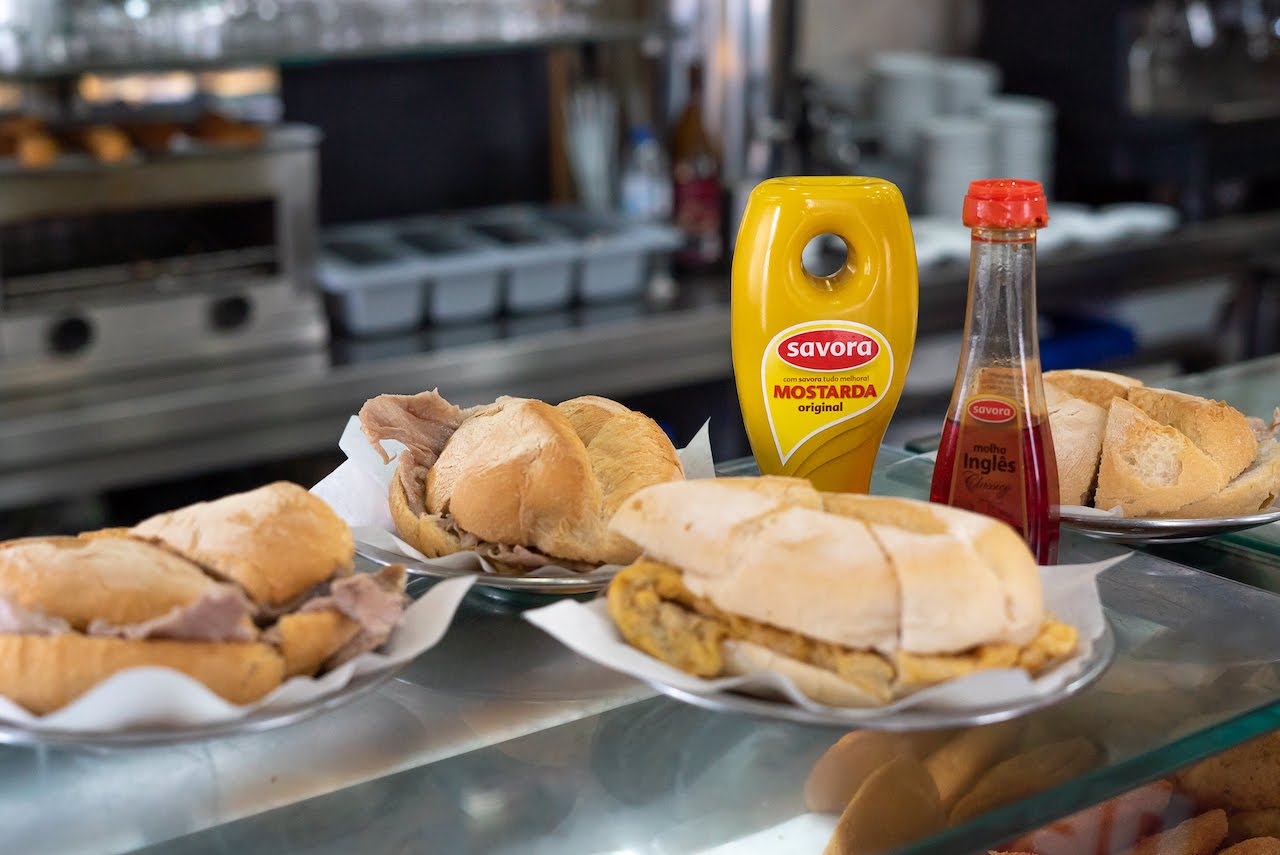 Bifana sandwiches on a restaurant coutner with mustard and other sauces