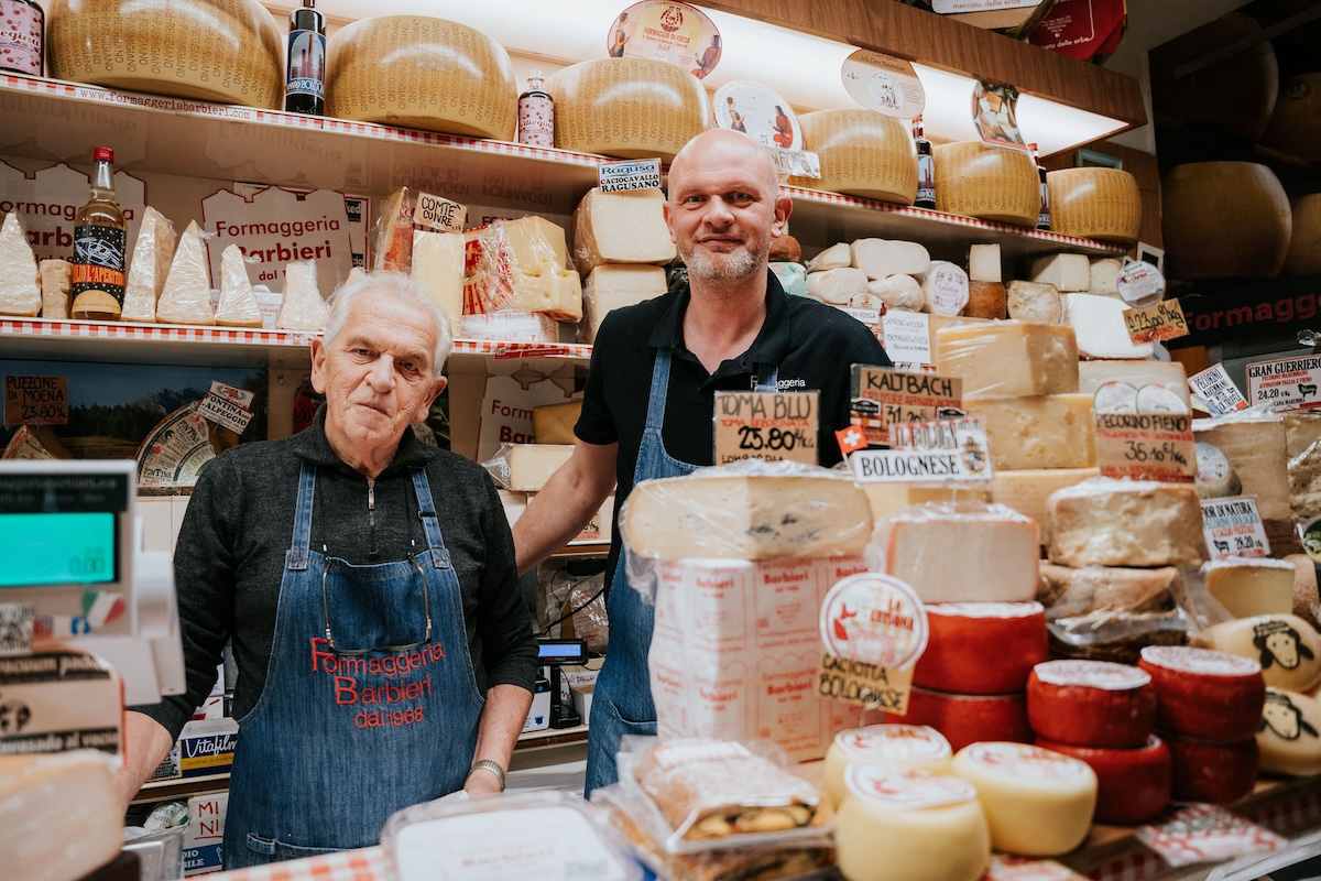 two older gentlemen working in food stand filled with different cheeses