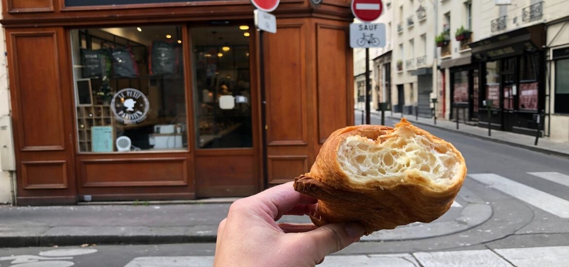 Hand holding croissant up with bite on street in front of a bakery in Paris