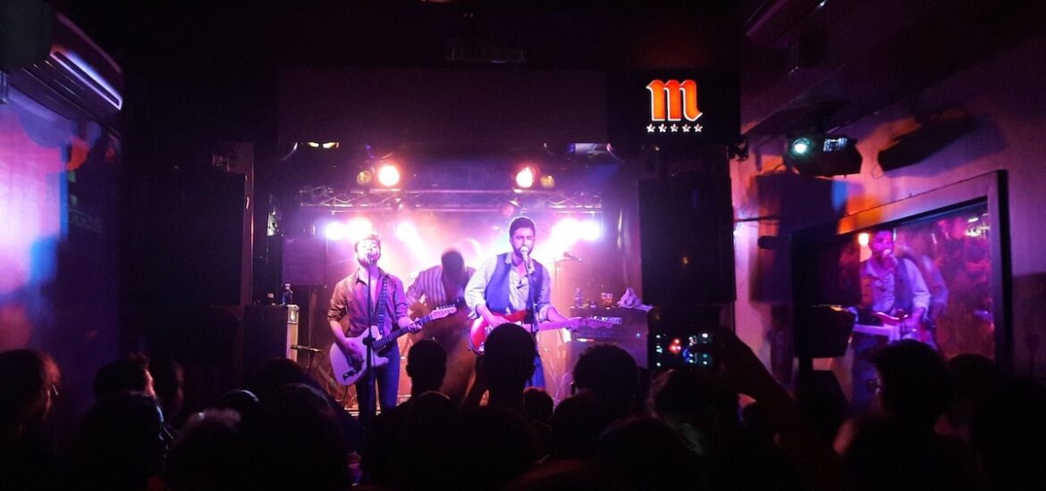 A rock band plays a concert inside a small venue with purple and white lighting illuminating the stage.