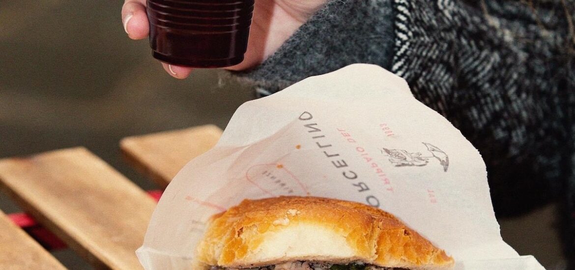 Lampredotto (tripe sandwich) in paper wrapping on a wooden table with a person's hand holding a plastic cup of red wine above