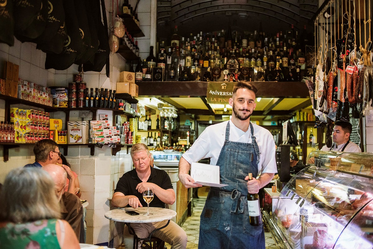 Interior of a wine bar in Barcelona with lots of bottles of wine above the heads of the patrons and a waiter who has a bottle of wine in hand