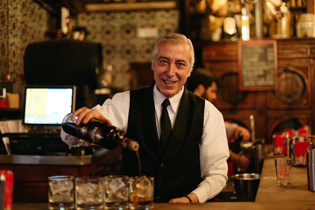 Barman or waiter behind counter at old historic wooden bar pouring vermouth into 4 clear glasses with ice