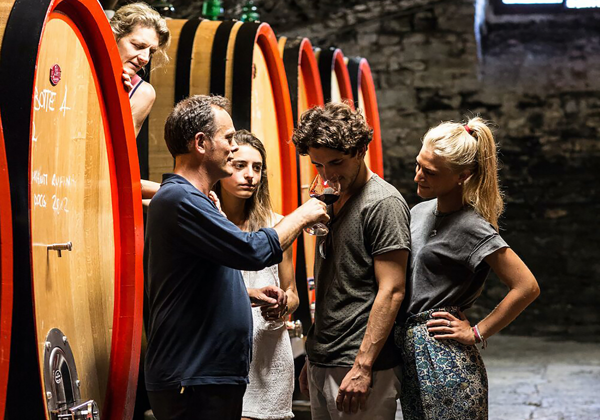 A group of people taking part in a wine tasting in front of a row of barrels