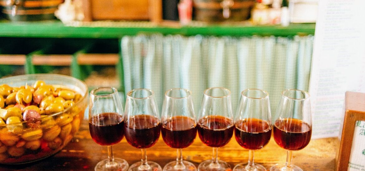 Six glasses of brown amontillado sherry wine lined up on a wooden bar top.