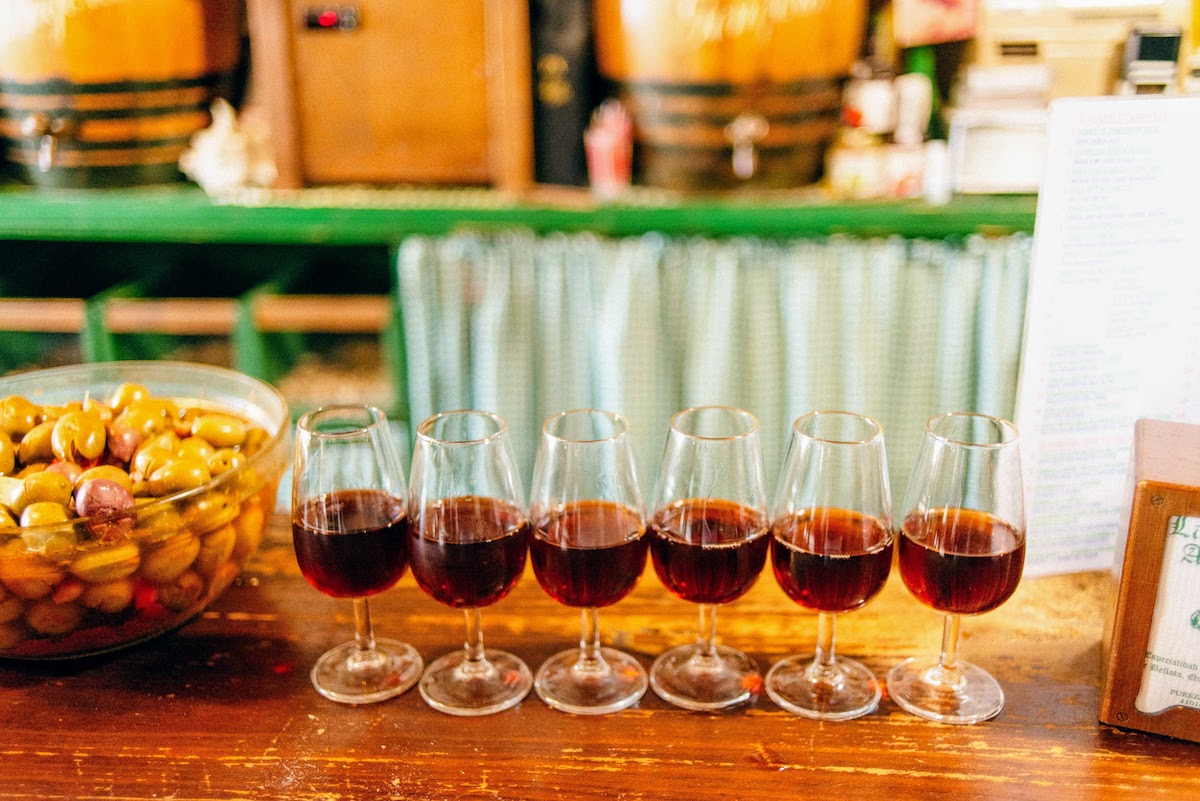 Six glasses of brown amontillado sherry wine lined up on a wooden bar top.