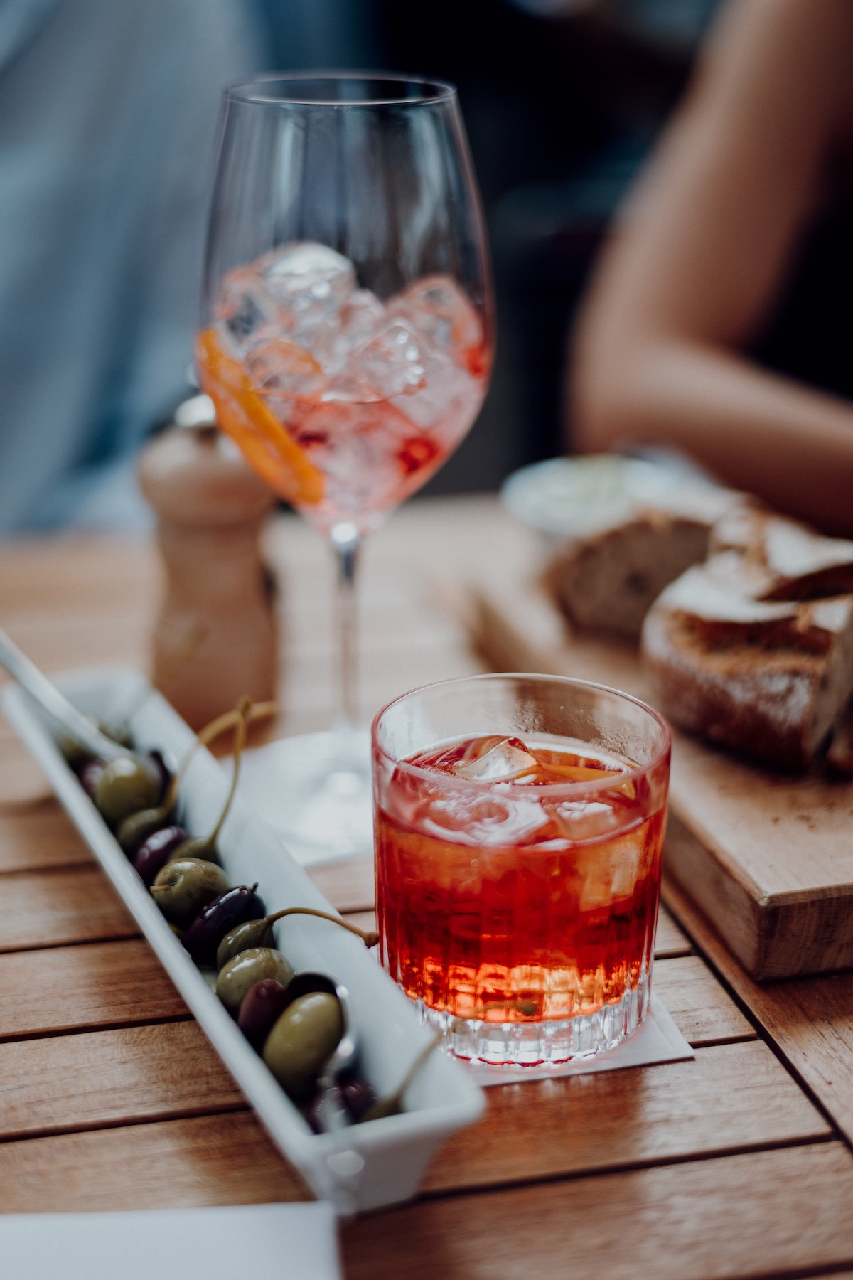 Reddish brown cocktail in a rocks glass beside a narrow dish of olives and a wine glass with ice cubes in the background