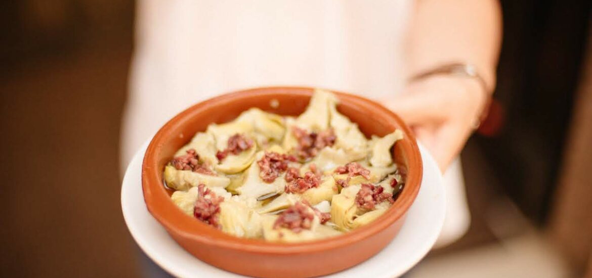 Person's hand holding a round clay dish of artichoke hearts sprinkled with diced cured ham.