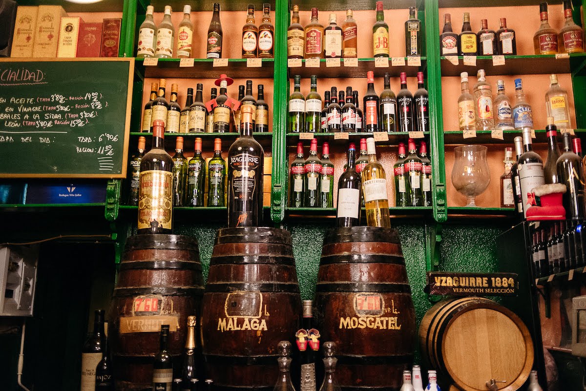 Interior of an old-fashioned Spanish bar with green wooden paneling, shelves full of bottles, and dark wood wine barrels.