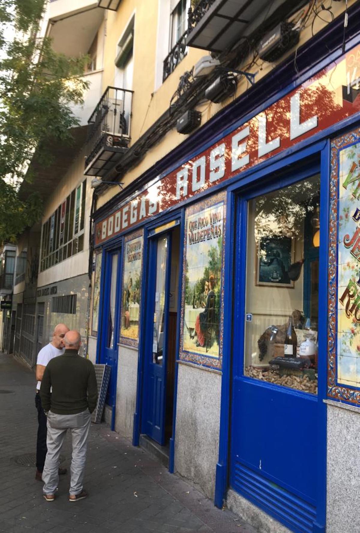 Two men talking outside of a bar with blue window paneling and old-fashioned tiled advertisements.