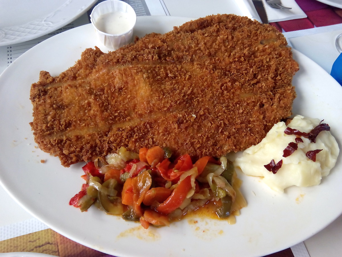 Large piece of fried meat on a white plate next to fried peppers and mashed potatoes