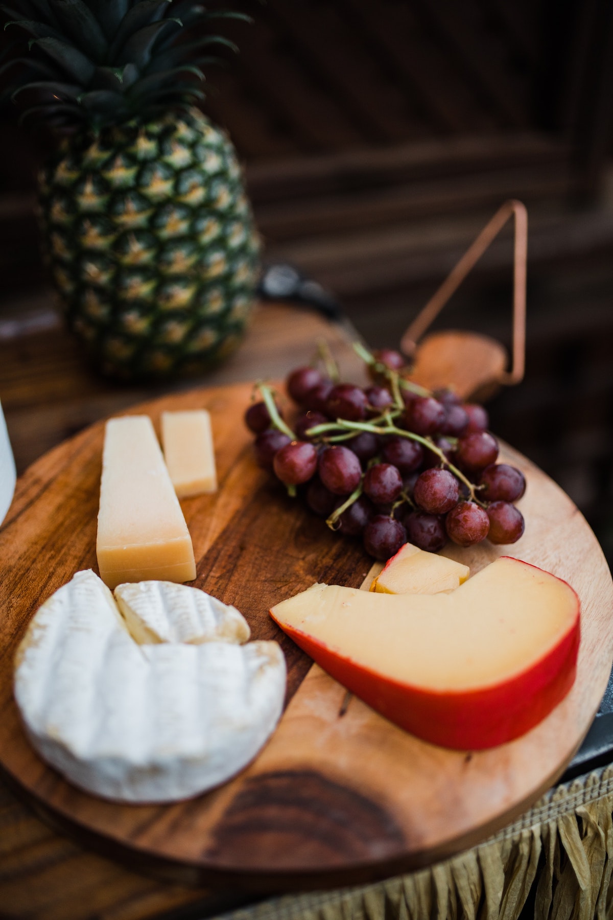 Three types of sliced cheese and a bunch of red grapes on a round wooden board with a pineapple visible in the background.