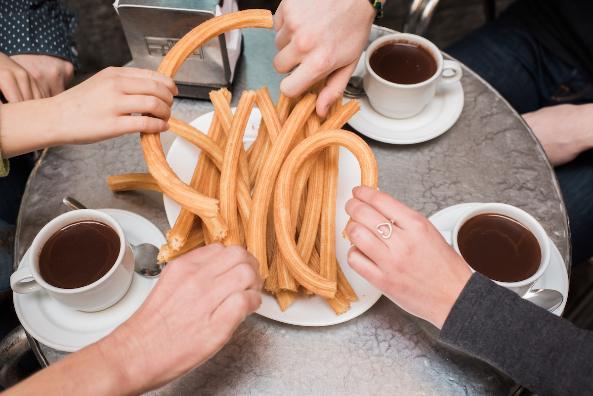 People grabbing churros off of a plate in the center of a table, with three mugs of hot chocolate nearby