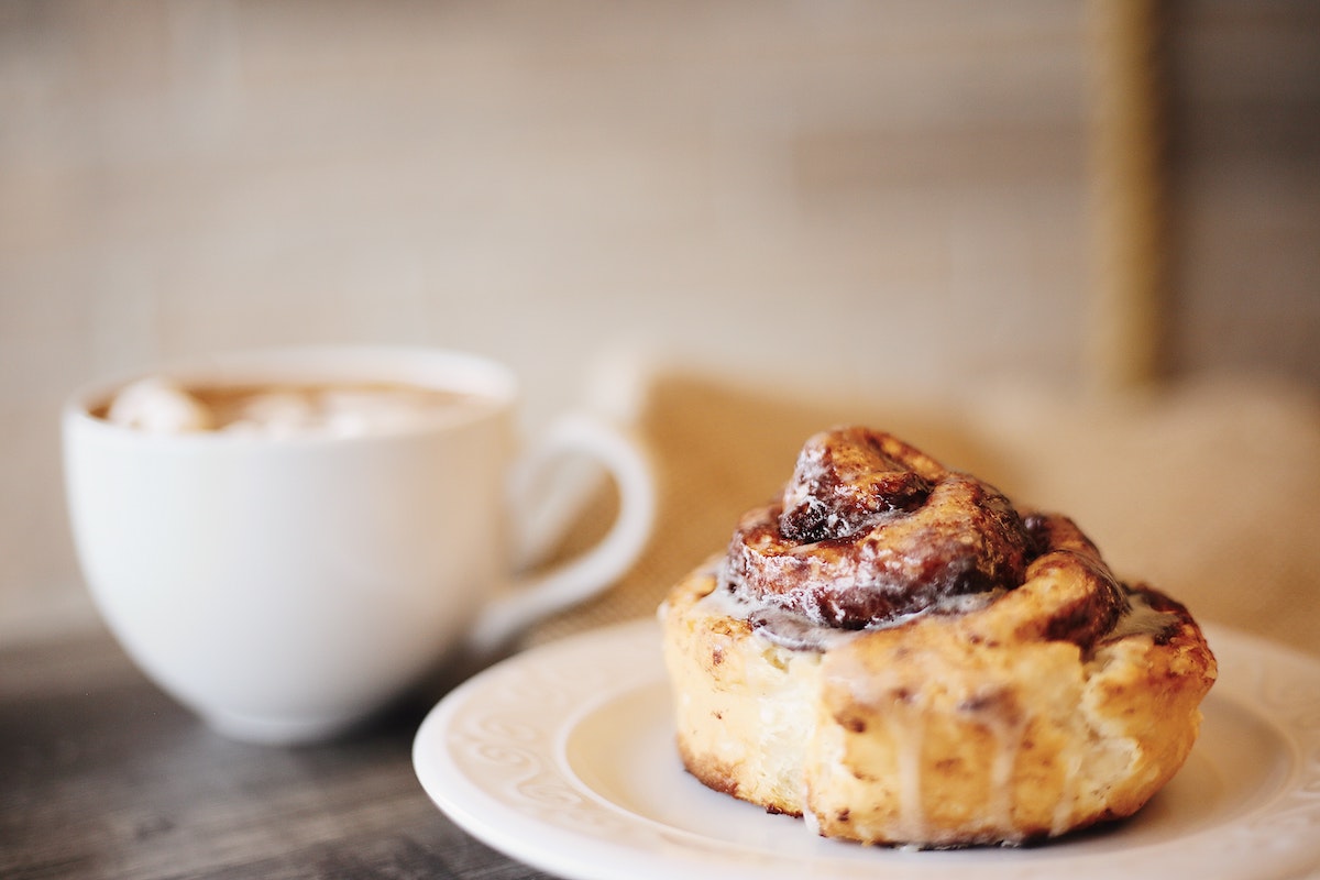 Cinnamon roll on a white plate next to a white cup of coffee.