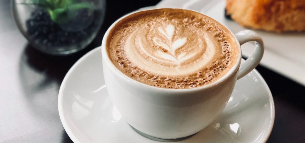 Two lattes in white mugs on a table beside a small potted plant and a pastry on a white plate.