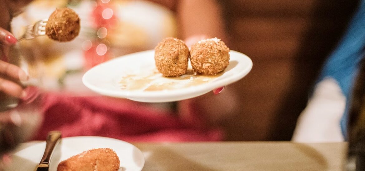 A person using a fork to take a fried croquette off a white plate.