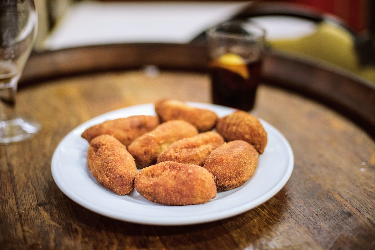 Several croquettes on a white plate, with a glass of vermouth visible in the background.