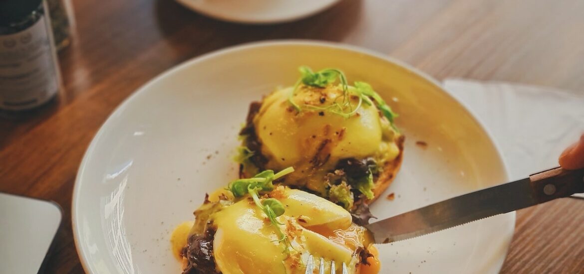 Person cutting into eggs Benedict with a cup of coffee on the table in the background