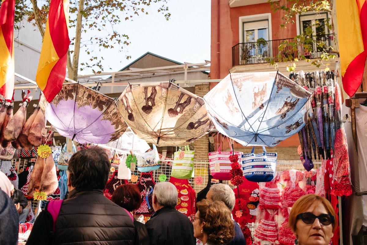 Stall at an open-air flea market selling umbrellas, tote bags, girls' dresses, and aprons.