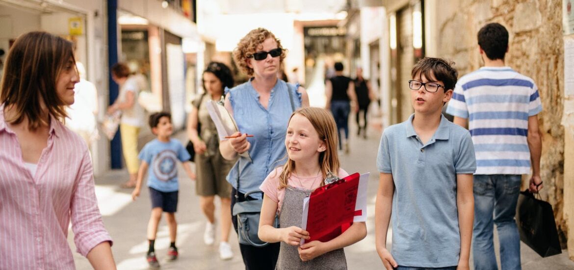 A little girl carrying a clipboard while walking through a busy city with a young boy and two women