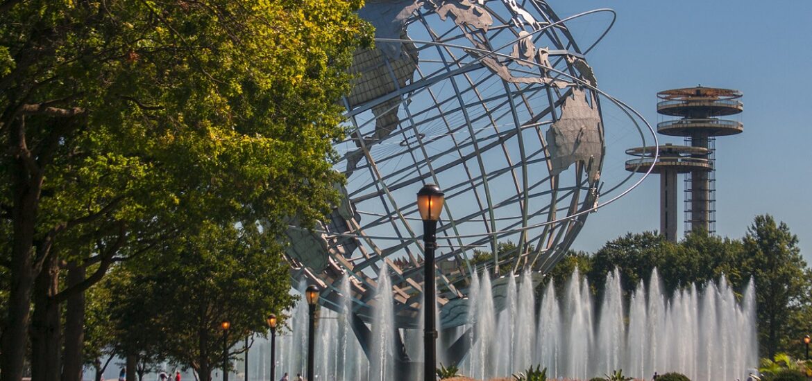 Huge silver sphere shaped like the earth emerges from behind trees and water fountains in a park in new york