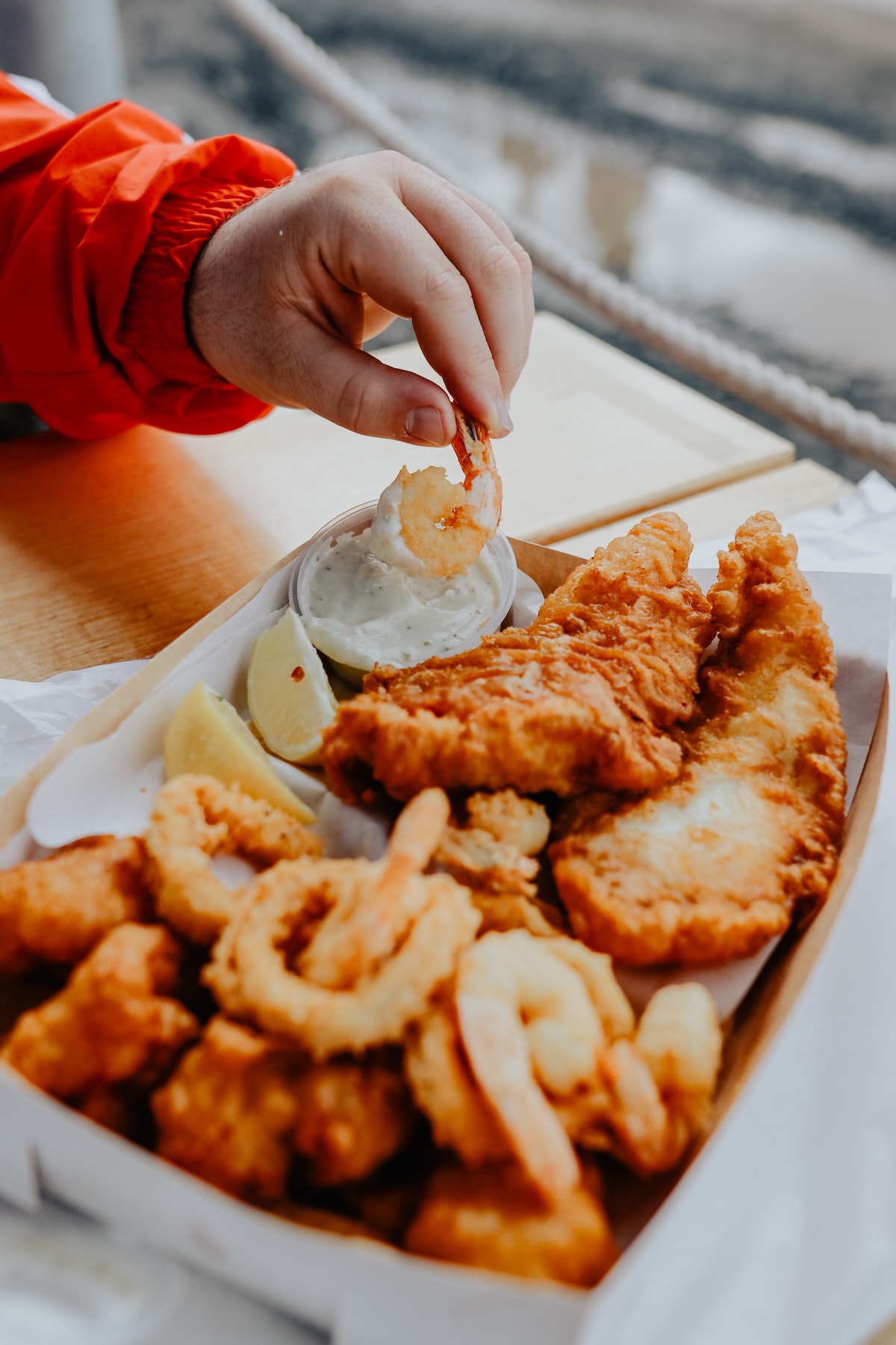 A person eating fried fish out of a box and dipping a piece of shrimp into white sauce