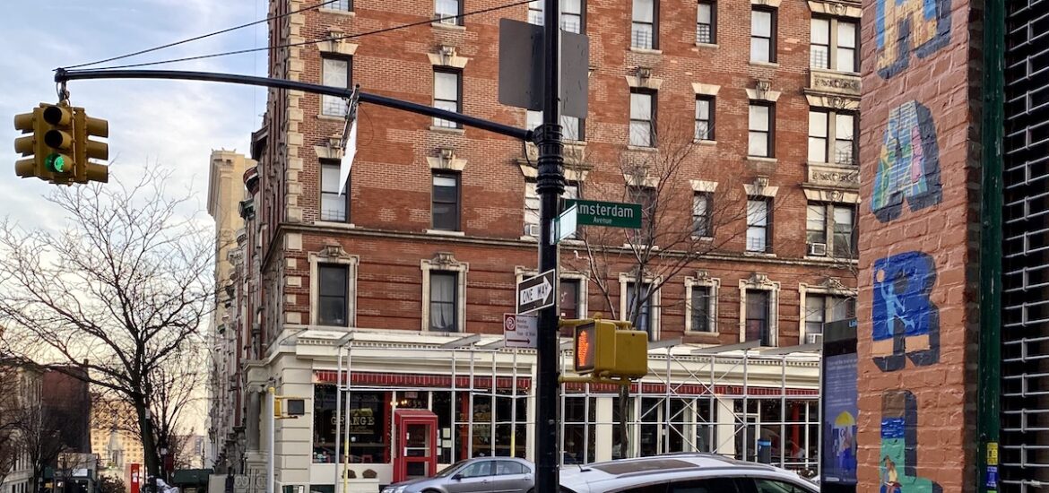 Intersection in an urban area with a multi-story brick building in the background and the word "Harlem" painted onto the building in the foreground.