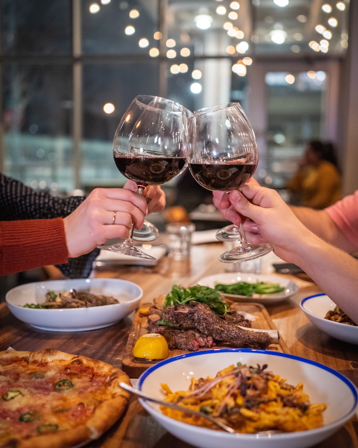 People clinking glasses of red wine above a table full of pizza, pasta, and steak