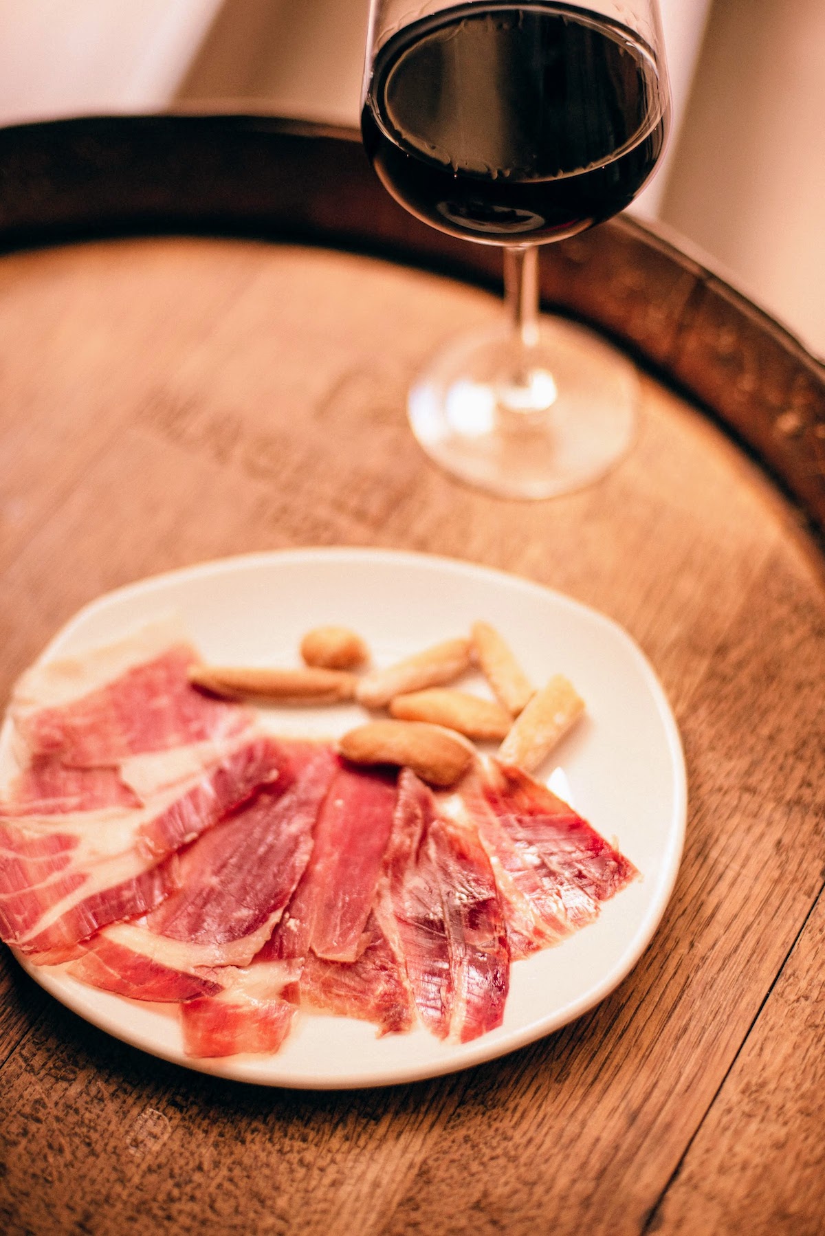 Cured ham and breadstick crackers on a white plate on top of a wooden barrel with a glass of red wine