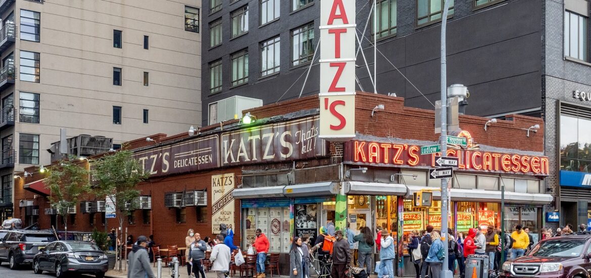 A busy street scene with people going into a restaurant and deli with a large neon sign