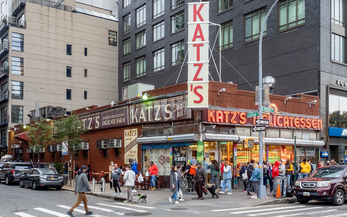 A busy street scene with people going into a restaurant and deli with a large neon sign