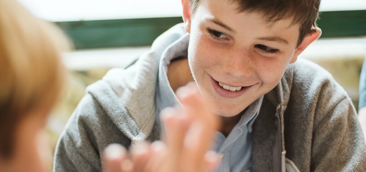 A boy smiling while drinking a mug of hot chocolate while looking at another boy seated at the same table just out of focus