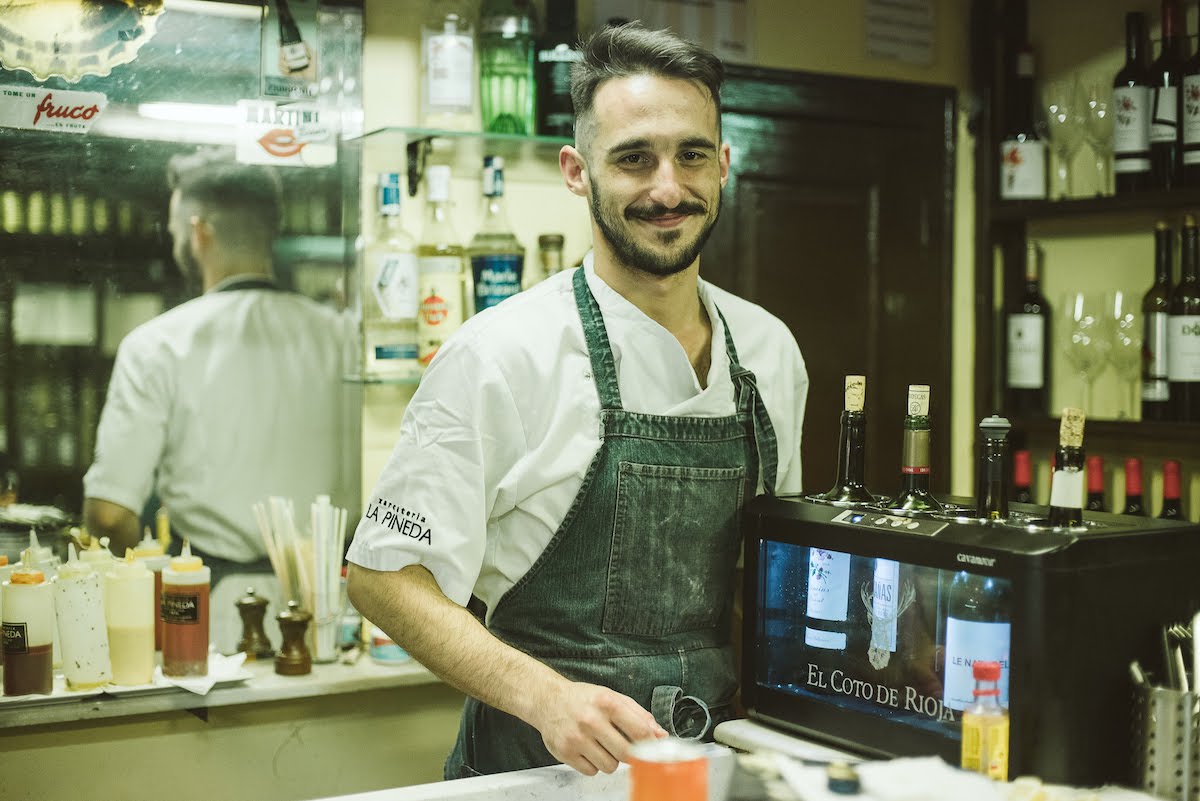 A man standing behind a bar beside a case of wine bottles.